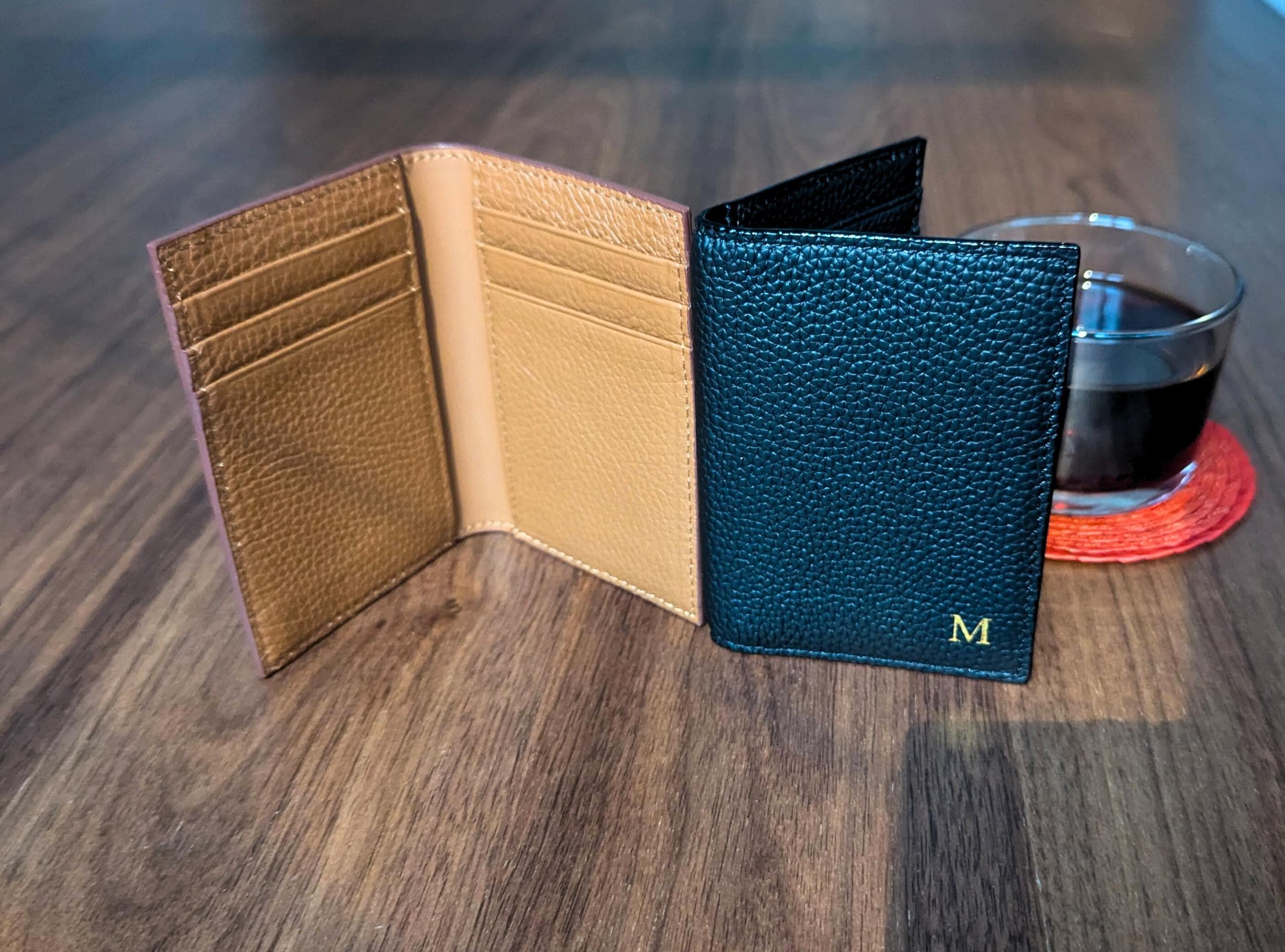 Two leather wallets, one beige and one blue with a visible 'M', on a wooden surface with a glass of dark liquid in the background.