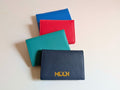 Four leather card holders in blue, red, green, and navy on a light beige background showing custom logo embossing. 