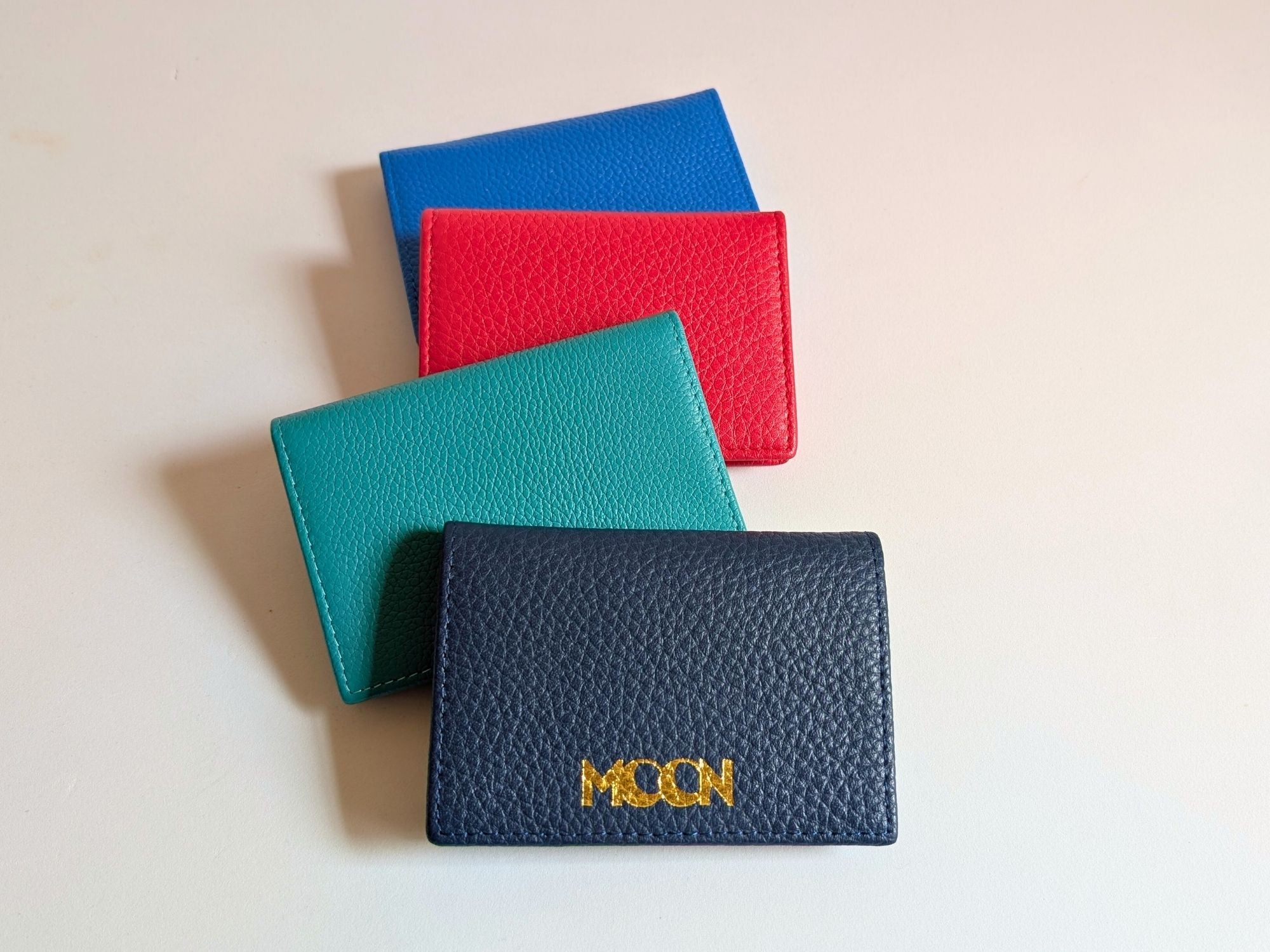 Four leather card holders in blue, red, green, and navy on a light beige background showing custom logo embossing. 