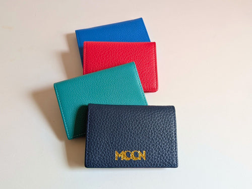 Four leather card holders in blue, red, green, and navy on a light beige background showing custom logo embossing. 