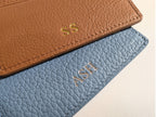 Close up of two leather card holders, one brown and one blue, with gold initials on a light beige background.