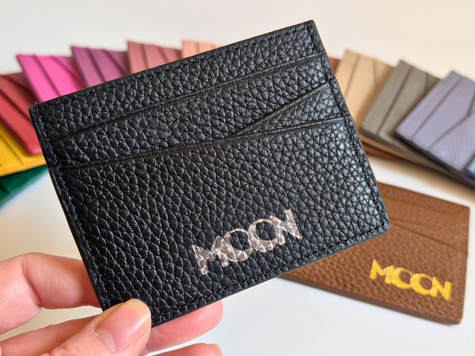 Black textured cardholder with custom branding held by a hand, with other color options in the background showing gold or silver emboss.