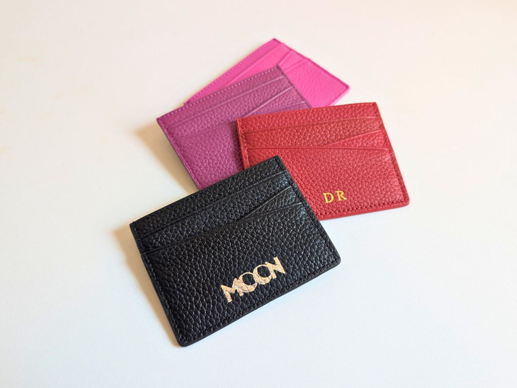 Black, red, and pink leather card holders on a light background displaying the custom logo in silver emboss and personalised initials in gold foil. 