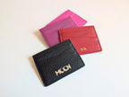 Black, red, and pink leather card holders on a light background displaying the custom logo in silver emboss and personalised initials in gold foil. 