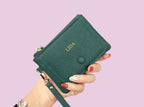Green wallet with embossed name held by a hand against a light purple background