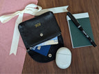 Black leather wallet with personalised gold monogram, airpods, notebook, and pen on a wooden surface.