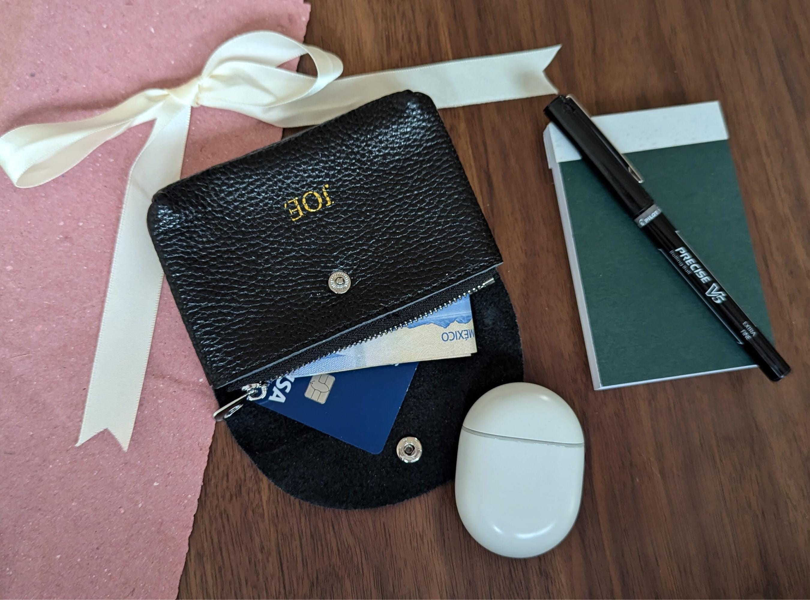 Black leather wallet with personalised gold monogram, airpods, notebook, and pen on a wooden surface.
