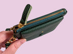 Green wallet with gold zipper held by a hand on a pink background