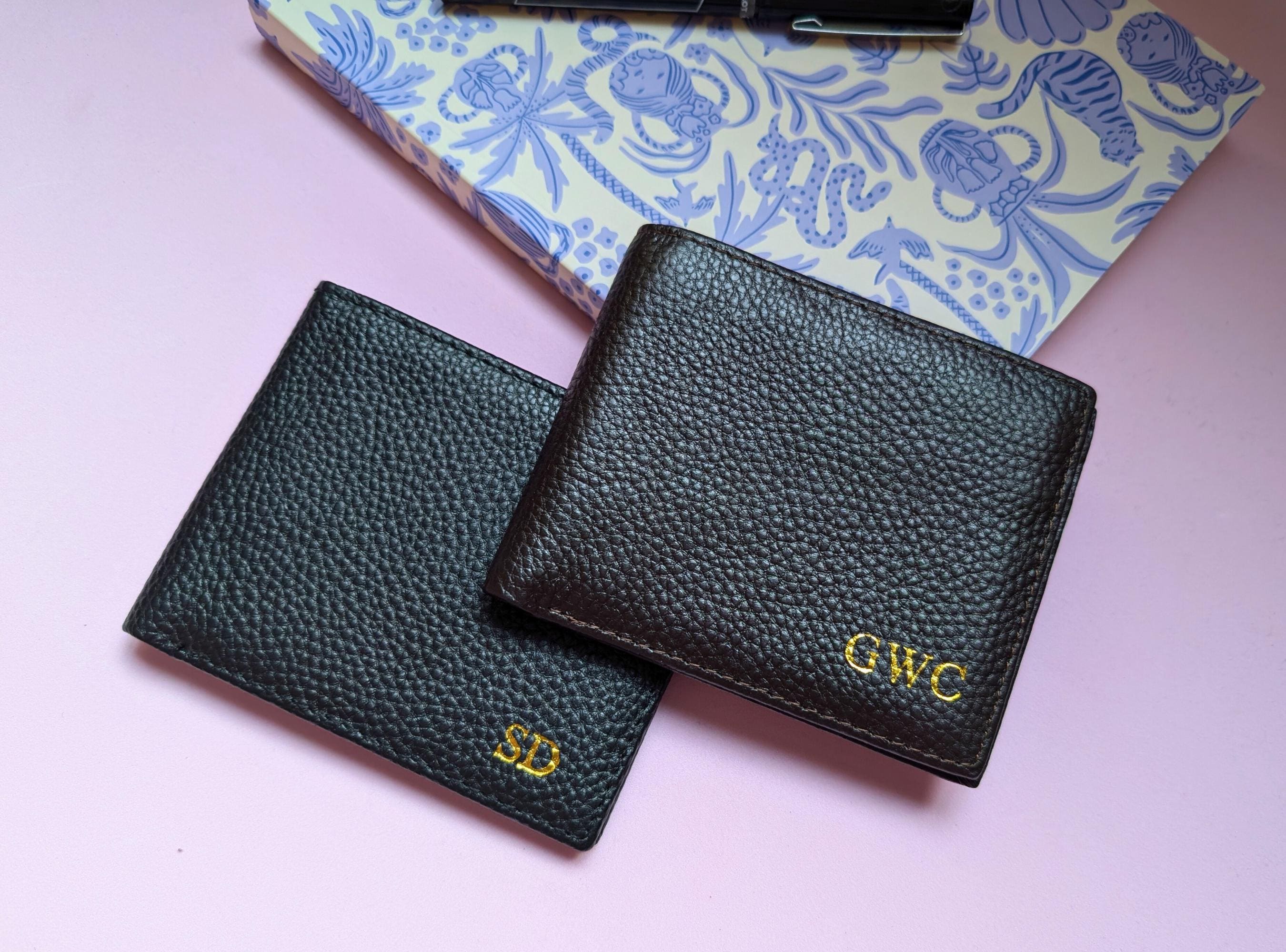 A black and chocolate leather wallets with initials on a pink background.
