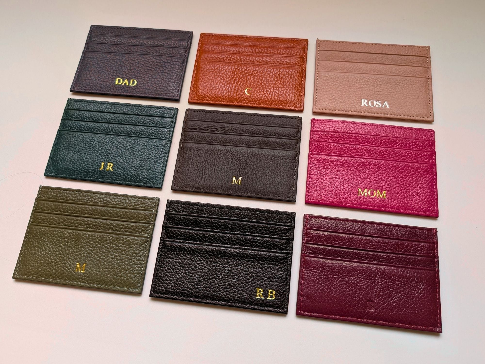Collection of leather card holders in various colors with initials engraved on a light gray background.