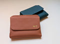 Brown leather wallet with gold initials and teal wallet on a light beige background
