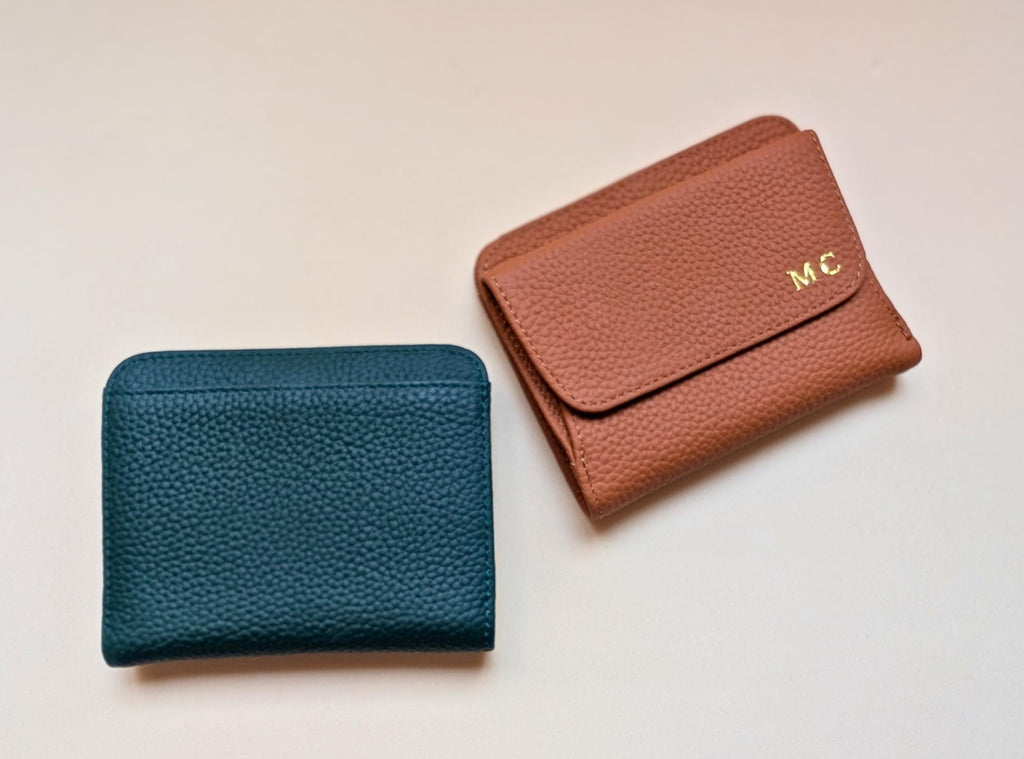 Two leather wallets, one blue and one brown with custom initials, on a light beige background.