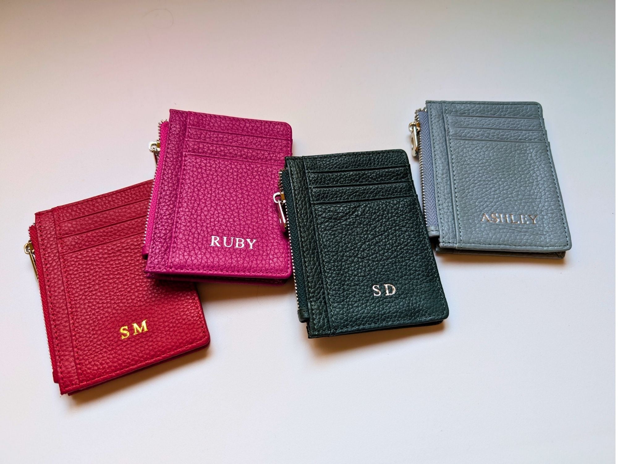 Four leather card holders in red, pink, black, and gray with initials on a light background